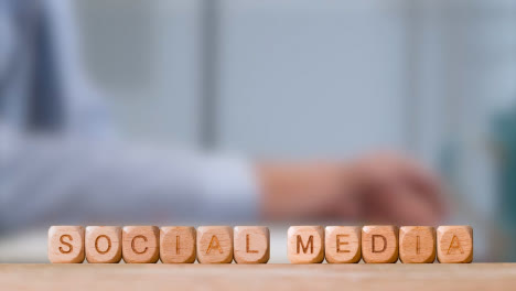 Business-Concept-Wooden-Letter-Cubes-Or-Dice-Spelling-Social-Media-With-Office-Person-Working-At-Laptop-In-Background