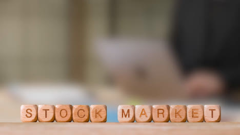 Business-Concept-Wooden-Letter-Cubes-Or-Dice-Spelling-Stock-Market-With-Office-Person-Working-At-Laptop-In-Background