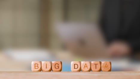 Business-Concept-Wooden-Letter-Cubes-Or-Dice-Spelling-Big-Data-With-Office-Person-Working-At-Laptop-In-Background