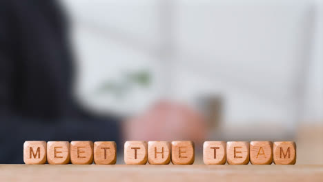 Business-Concept-Wooden-Letter-Cubes-Or-Dice-Spelling-Meet-The-Team-With-Office-Person-Using-Mobile-Phone-In-Background
