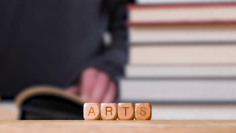 Education-Concept-Shot-With-Wooden-Letter-Cubes-Or-Dice-Spelling-Arts-With-Person-Reading-Book-In-Library