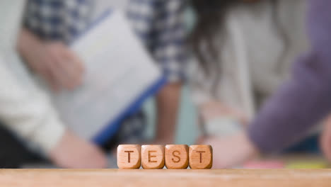 Education-Concept-With-Wooden-Letter-Cubes-Or-Dice-Spelling-Test-With-Students-Meeting-In-Background