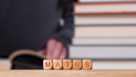 Education-Concept-Shot-With-Wooden-Letter-Cubes-Or-Dice-Spelling-Maths-With-Person-Reading-Book-In-Library