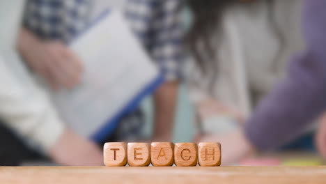 Education-Concept-With-Wooden-Letter-Cubes-Or-Dice-Spelling-Teach-With-Students-Meeting-In-Background