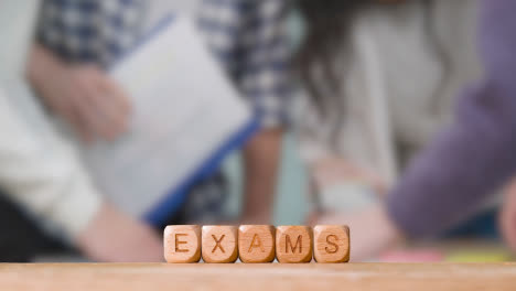 Education-Concept-With-Wooden-Letter-Cubes-Or-Dice-Spelling-Exams-With-Students-Meeting-In-Background