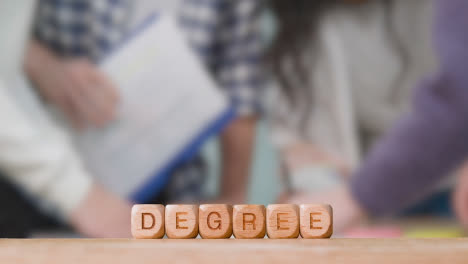 Education-Concept-With-Wooden-Letter-Cubes-Or-Dice-Spelling-Degree-With-Students-Meeting-In-Background