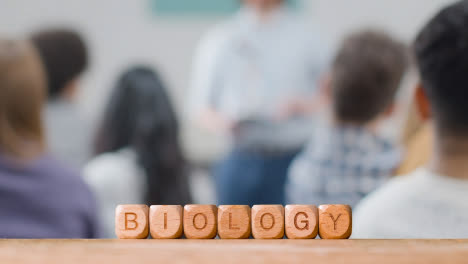 Education-Concept-With-Wooden-Letter-Cubes-Or-Dice-Spelling-Biology-With-Student-Lecture-In-Background