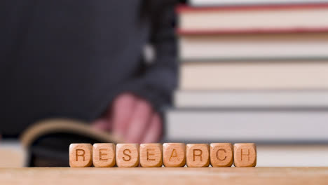 Education-Concept-Shot-With-Wooden-Letter-Cubes-Or-Dice-Spelling-Research-With-Person-Reading-Book-In-Library