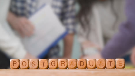 Education-Concept-With-Wooden-Letter-Cubes-Or-Dice-Spelling-Postgraduate-With-Students-Meeting-In-Background