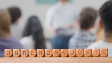 Education-Concept-With-Wooden-Letter-Cubes-Or-Dice-Spelling-Undergraduate-With-Students-Meeting-In-Background