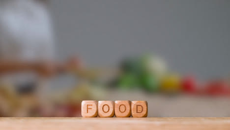 Concepto-Con-Cubos-De-Letras-De-Madera-O-Dados-Que-Deletrean-Comida-Contra-El-Fondo-De-Una-Mujer-Cortando-Ingredientes