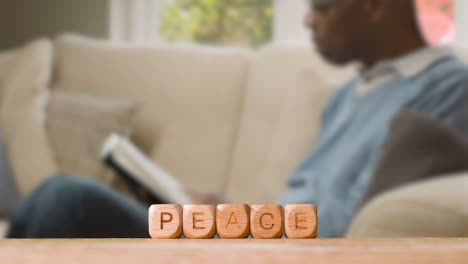 Concept-With-Wooden-Letter-Cubes-Or-Dice-Spelling-Peace-Against-Background-Of-Man-Sitting-On-Sofa-Reading-Book