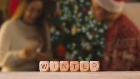 Konzept-Mit-Hölzernen-Buchstabenwürfeln-Oder-Würfeln,-Die-Winter-Vor-Dem-Hintergrund-Eines-Paares-Buchstabieren,-Das-Zu-Weihnachten-Geschenke-Austauscht