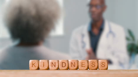 Concept-With-Wooden-Letter-Cubes-Or-Dice-Spelling-Kindness-Against-Background-Of-Doctor-Talking-To-Patient-In-Hospital