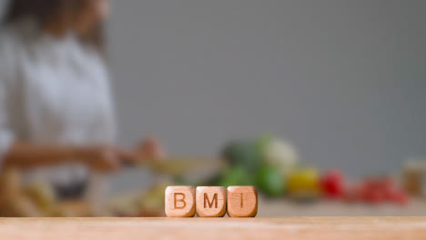 Medical-Concept-With-Wooden-Letter-Cubes-Or-Dice-Spelling-BMI-Against-Background-Of-Woman-Chopping-Ingredients