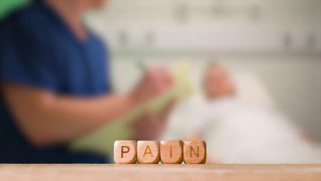 Medical-Concept-With-Wooden-Letter-Cubes-Or-Dice-Spelling-Pain-Against-Background-Of-Nurse-Talking-To-Patient-In-Hospital-Bed