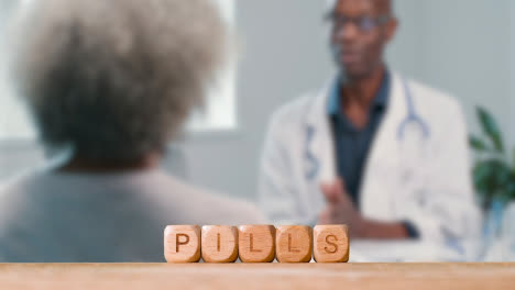 Medical-Concept-With-Wooden-Letter-Cubes-Or-Dice-Spelling-Pills-Against-Background-Of-Doctor-Talking-To-Patient-In-Hospital