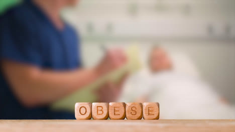 Medical-Concept-With-Wooden-Letter-Cubes-Or-Dice-Spelling-Obese-Against-Background-Of-Nurse-Talking-To-Patient-In-Hospital-Bed