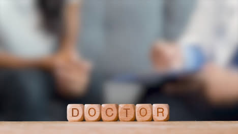 Medical-Concept-With-Wooden-Letter-Cubes-Or-Dice-Spelling-Doctor-Against-Background-Of-GP-Talking-To-Patient-In-Hospital
