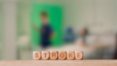 Medical-Concept-With-Wooden-Letter-Cubes-Or-Dice-Spelling-Stroke-Against-Background-Of-Nurse-Talking-To-Patient-In-Hospital-Bed
