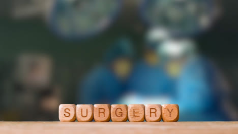 Medical-Concept-With-Wooden-Letter-Cubes-Or-Dice-Spelling-Surgery-Against-Background-Of-Operation-In-Theatre