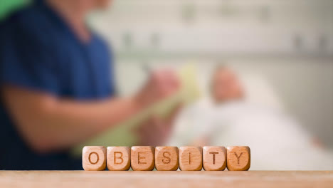 Medical-Concept-With-Wooden-Letter-Cubes-Or-Dice-Spelling-Obesity-Against-Background-Of-Nurse-Talking-To-Patient-In-Hospital-Bed
