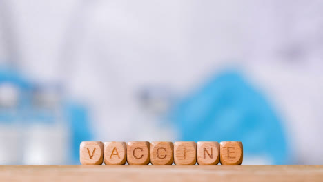 Medical-Concept-With-Wooden-Letter-Cubes-Or-Dice-Spelling-Vaccine-Against-Background-Of-Scientist-Working-In-Laboratory