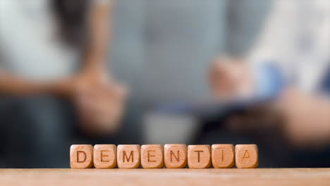 Medical-Concept-With-Wooden-Letter-Cubes-Or-Dice-Spelling-Dementia-Against-Background-Of-Doctor-Talking-To-Patient