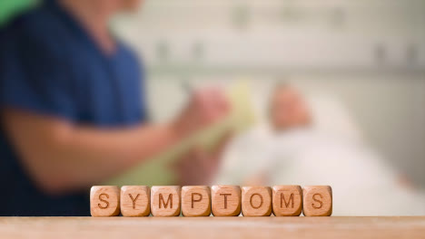 Medical-Concept-With-Wooden-Letter-Cubes-Or-Dice-Spelling-Symptoms-Against-Background-Of-Nurse-Talking-To-Patient-In-Hospital-Bed