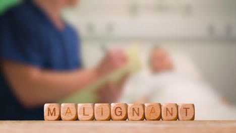 Medical-Concept-With-Wooden-Letter-Cubes-Or-Dice-Spelling-Malignant-Against-Background-Of-Nurse-Talking-To-Patient-In-Hospital-Bed