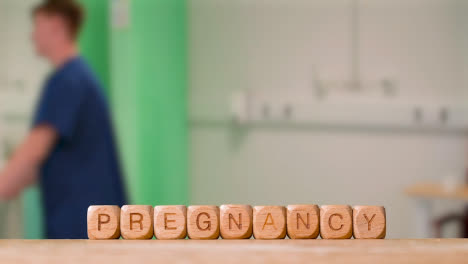 Medical-Concept-With-Wooden-Letter-Cubes-Or-Dice-Spelling-Pregnancy-Against-Background-Of-Nurse-Wheeling-Patient-In-Hospital-Bed