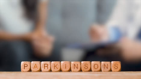 Medical-Concept-With-Wooden-Letter-Cubes-Or-Dice-Spelling-Parkinson's-Against-Background-Of-Doctor-Talking-To-Patient