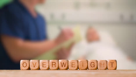Medical-Concept-With-Wooden-Letter-Cubes-Or-Dice-Spelling-Overweight-Against-Background-Of-Nurse-Talking-To-Patient-In-Hospital-Bed