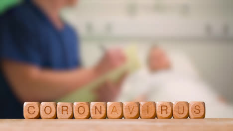 Medical-Concept-With-Wooden-Letter-Cubes-Or-Dice-Spelling-Coronavirus-Against-Background-Of-Nurse-Talking-To-Patient-In-Hospital-Bed