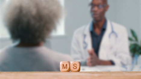 Medizinisches-Konzept-Mit-Holzbuchstabenwürfeln-Oder-Würfeln,-Die-Ms-Vor-Dem-Hintergrund-Des-Arztes-Im-Gespräch-Mit-Dem-Patienten-Buchstabieren