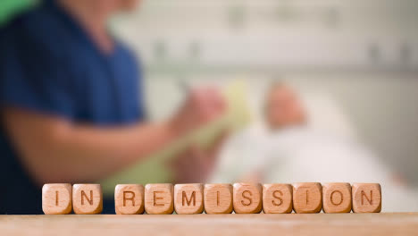 Medical-Concept-With-Wooden-Letter-Cubes-Or-Dice-Spelling-In-Remission-Against-Background-Of-Nurse-Talking-To-Patient-In-Hospital-Bed