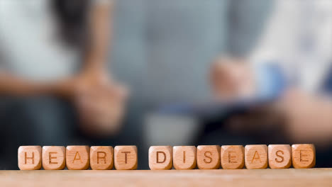 Medical-Concept-With-Wooden-Letter-Cubes-Or-Dice-Spelling-Heart-Disease-Against-Background-Of-Doctor-Talking-To-Patient