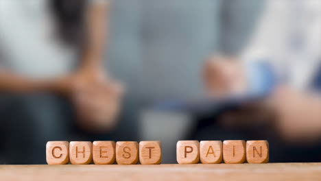 Medical-Concept-With-Wooden-Letter-Cubes-Or-Dice-Spelling-Chest-Pain-Against-Background-Of-Doctor-Talking-To-Patient