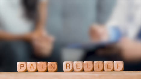 Medical-Concept-With-Wooden-Letter-Cubes-Or-Dice-Spelling-Pain-Relief-Against-Background-Of-Doctor-Talking-To-Patient