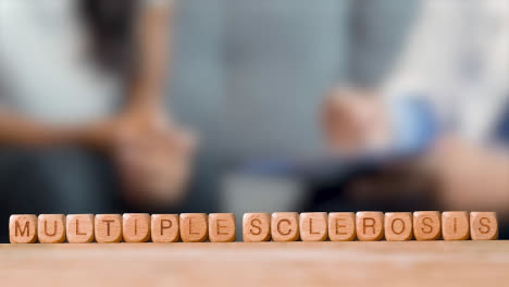 Medical-Concept-With-Wooden-Letter-Cubes-Or-Dice-Spelling-Multiple-Sclerosis-Against-Background-Of-Doctor-Talking-To-Patient