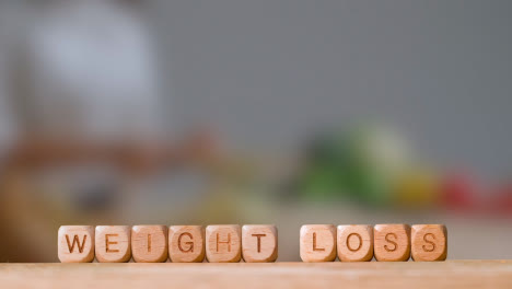 Medical-Concept-With-Wooden-Letter-Cubes-Or-Dice-Spelling-Weight-Loss-Against-Background-Of-Woman-Chopping-Ingredients