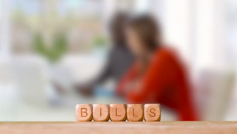 Concept-With-Wooden-Letter-Cubes-Or-Dice-Spelling-Bills-Against-Background-Of-Couple-Looking-At-Bills