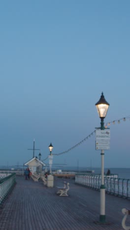 Vertikales-Video-Von-Menschen,-Die-In-Der-Dämmerung-Am-Penarth-Pier-In-Wales-Spazieren