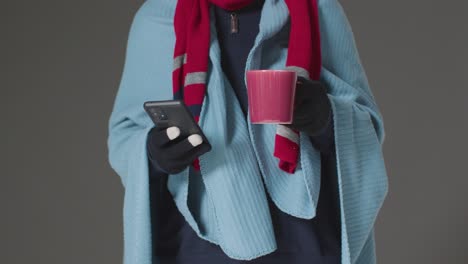 Close-Up-Of-Person-Wearing-Blanket-With-Hot-Drink-Using-Mobile-Phone-Trying-To-Keep-Warm-In-Energy-Crisis