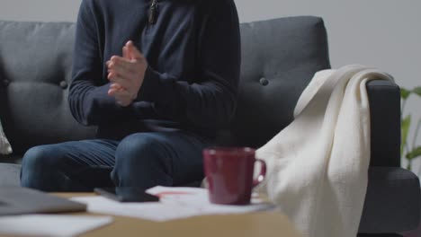 Person-Wearing-Blanket-With-Hot-Drink-Sitting-On-Sofa-At-Home-Trying-To-Keep-Warm-In-Energy-Crisis