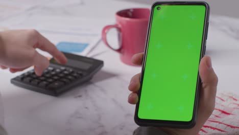 Close-Up-Of-Person-Holding-Green-Screen-Mobile-Phone-Looking-Through-Bills-In-Kitchen