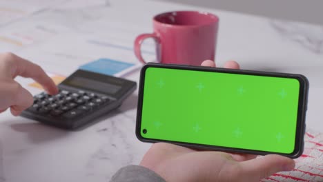 Primer-Plano-De-La-Persona-Que-Sostiene-Un-Teléfono-Móvil-De-Pantalla-Verde-Mirando-Las-Facturas-En-La-Cocina-1