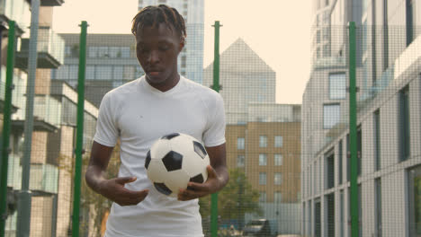 Portrait-Of-Young-Man-With-Football-Under-Arm-On-Artificial-Soccer-Pitch-In-Urban-City-Area-