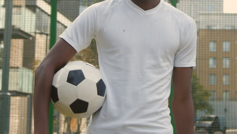 Young-Man-With-Football-Under-Arm-On-Artificial-Soccer-Pitch-In-Urban-City-Area-