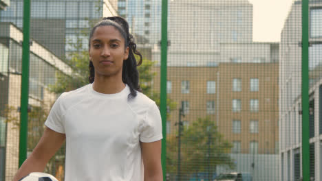 Portrait-Of-Young-Woman-Holding-Football-On-Artificial-Soccer-Pitch-In-Urban-City-Area-1